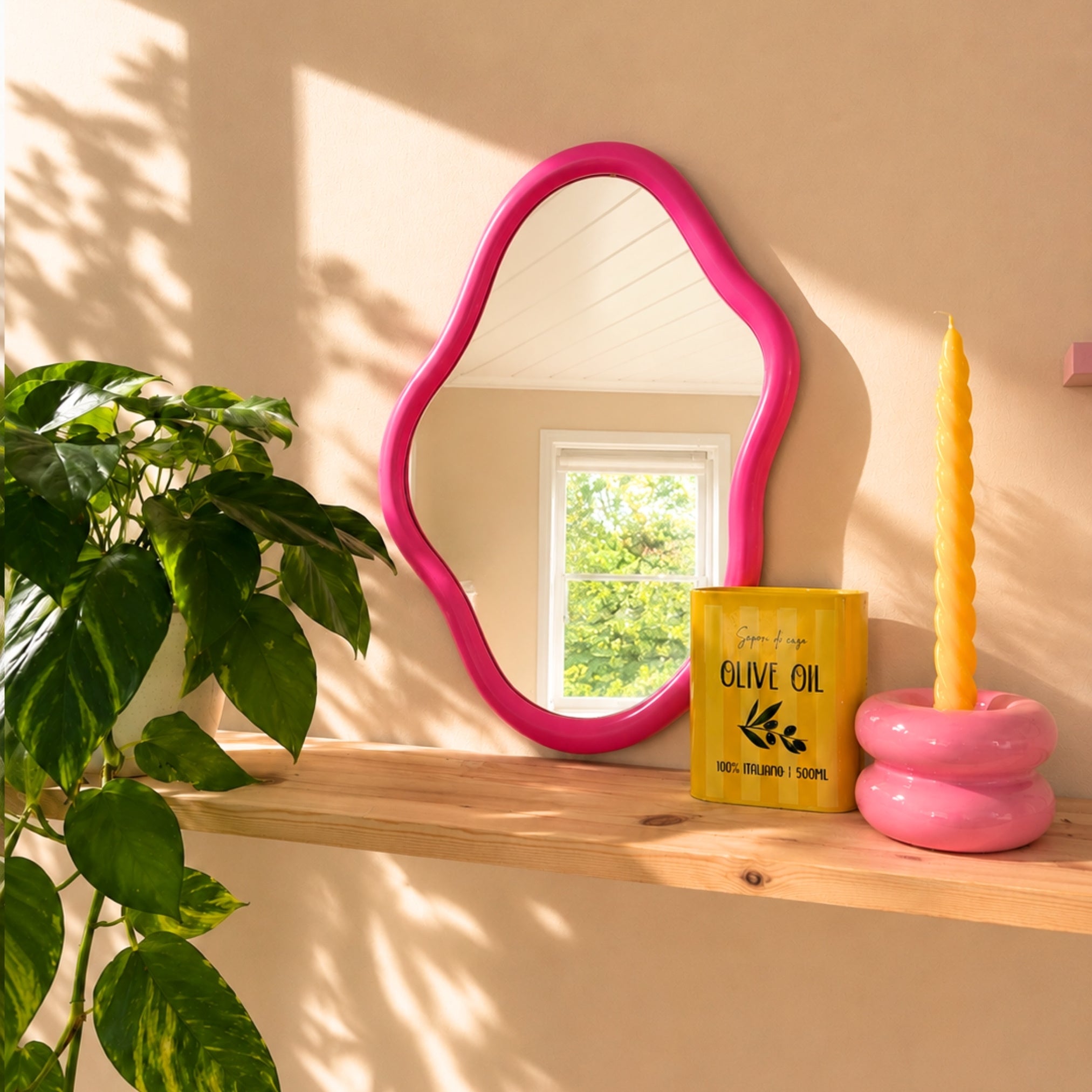 Rosa bølgete speil på trehylle med grønne planter, varm beige vegg og naturlig sollys.