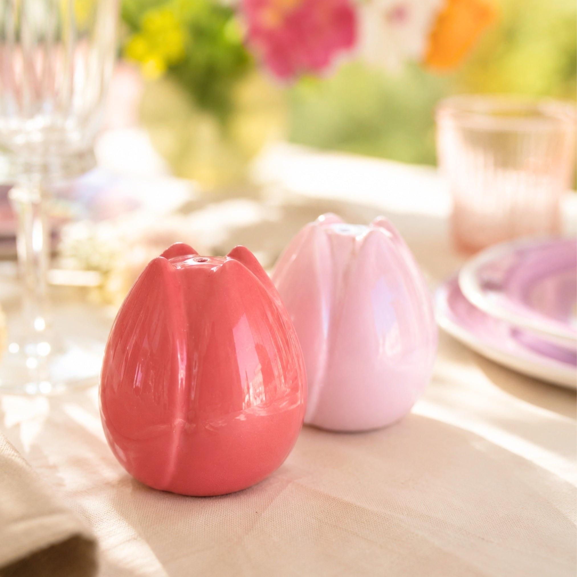 Blomsterformet salt- og pepperbøsser i rosa og korall på dekket bord med naturlig sollys og fargerik borddekking.
